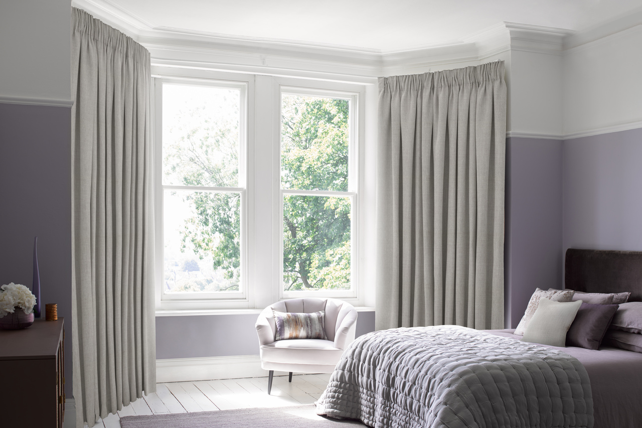 Elegant bedroom with soft grey pinch pleat curtains framing large windows, featuring a cosy armchair and a quilted bedspread