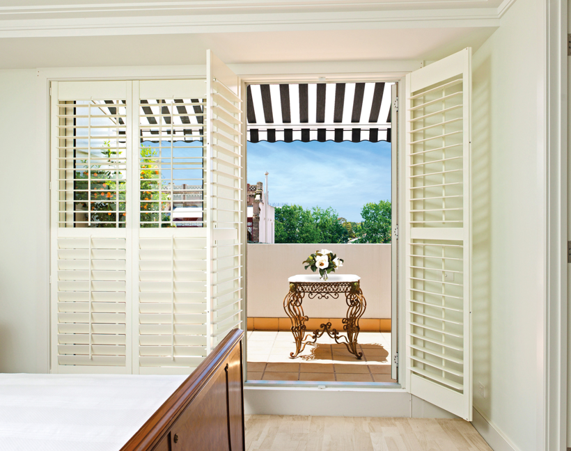 Balcony with a black and white striped awning providing shade, viewed from a bedroom through open white plantation shutters, creating a cosy and stylish outdoor retreat
