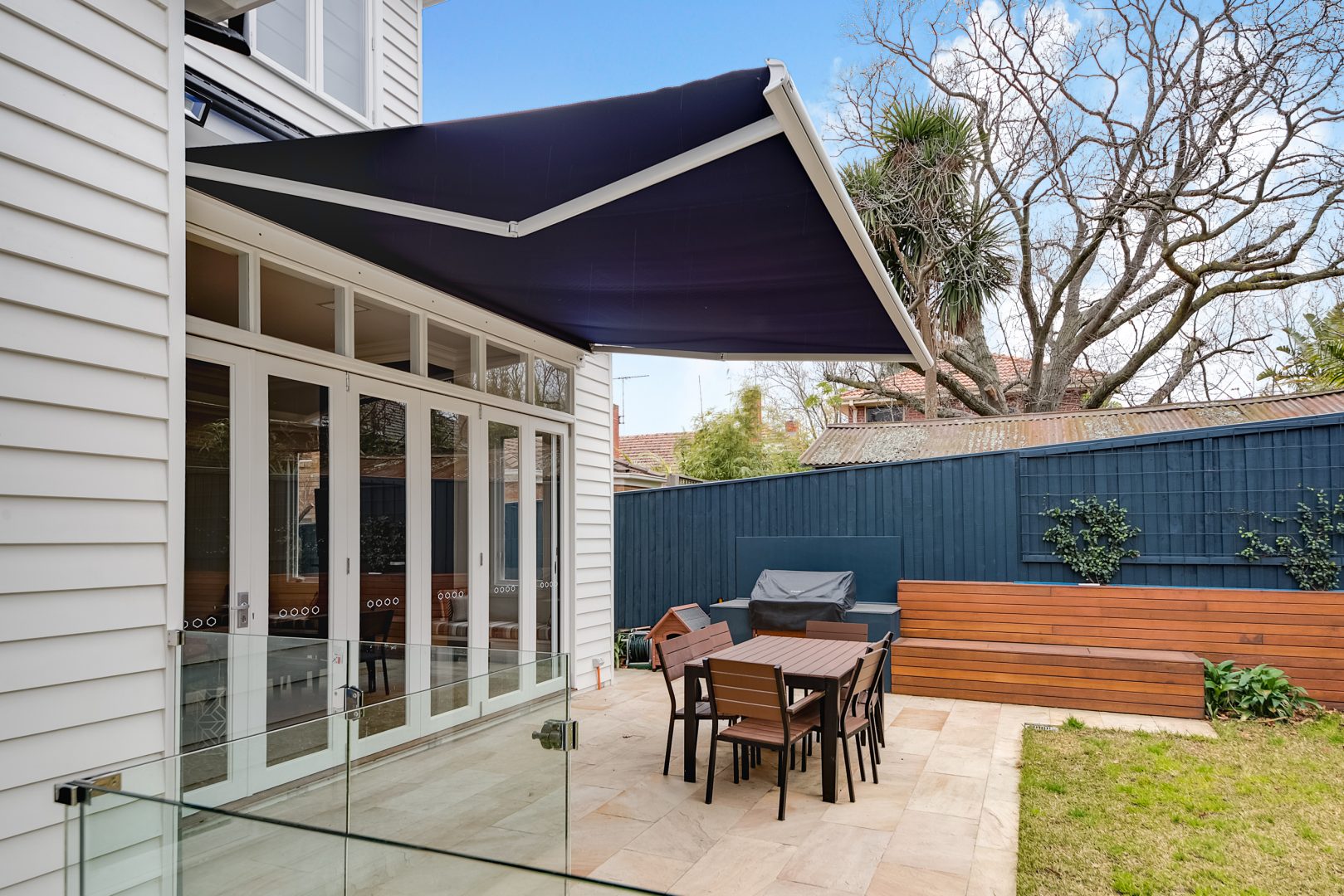 Navy and white folding arm owning up and open over an outdoor dining set and patio connected to a white paneled house