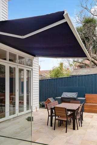 Navy and white folding arm owning up and open over an outdoor dining set and patio connected to a white paneled house