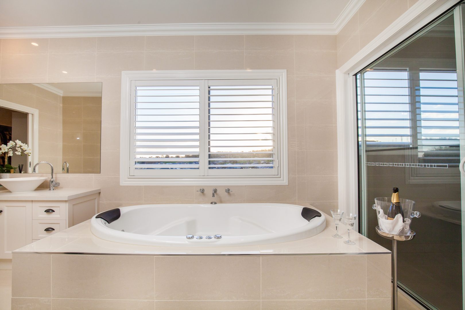 Luxurious bathroom featuring a large white bathtub, beige tiled walls, and a window with white plantation shutters, offering privacy and a serene view