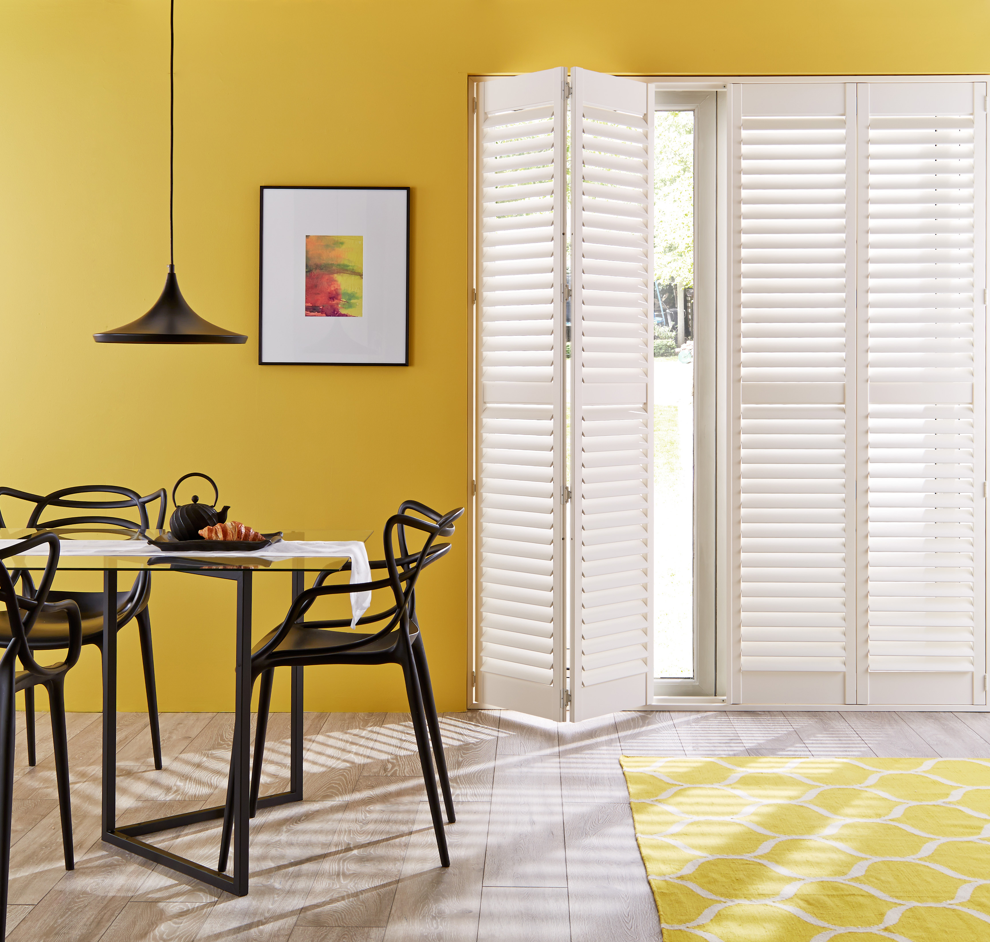 Bright dining area with white plantation shutters, yellow walls, a glass dining table with black chairs, and a yellow patterned rug, creating a cheerful and modern space