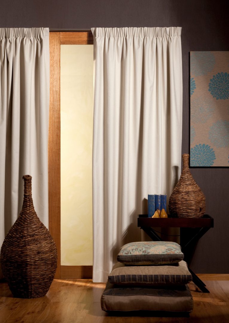 Living room with cream pinch-pleat curtains, wicker décor, and stacked cushions on a wooden floor, creating a warm, rustic atmosphere