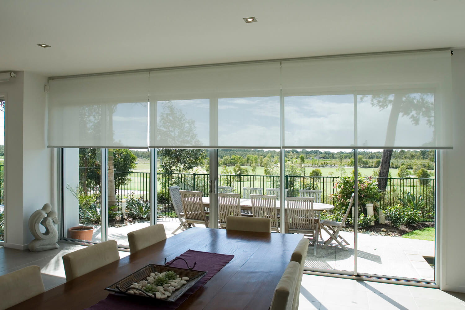 Dining room with large glass patio doors, featuring sheer roller blinds and a wooden dining table with neutral-toned chairs, overlooking a lush outdoor garden space