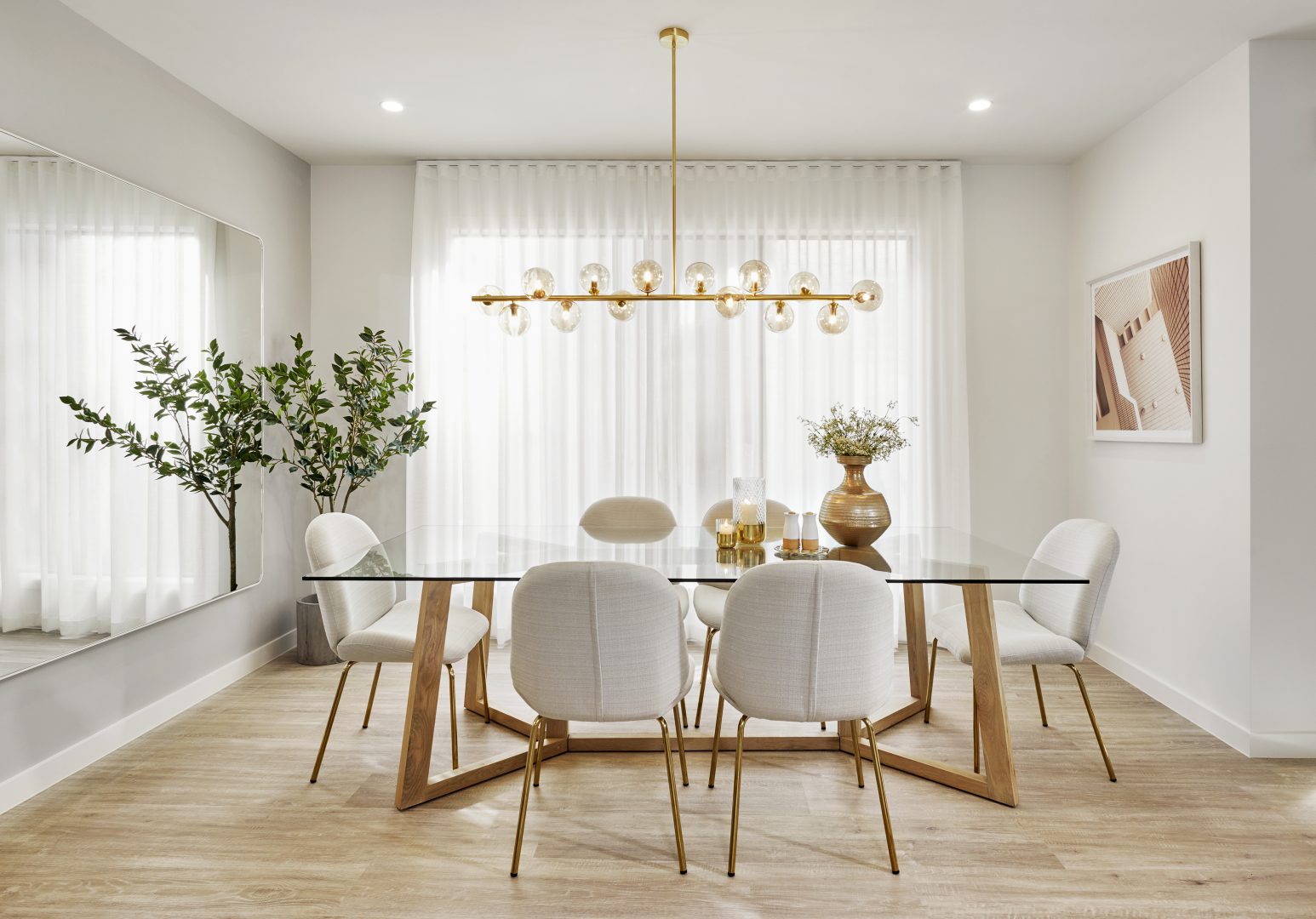 Modern dining room featuring sheer white curtains, a glass dining table with wooden legs, and upholstered chairs, complemented by a contemporary chandelier and decorative plants