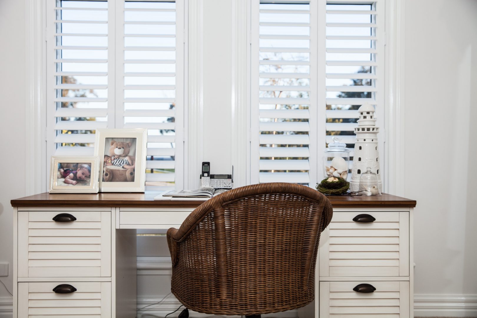 Office room with a brown wicker chair and cream and brown desk complimented by open white shutters