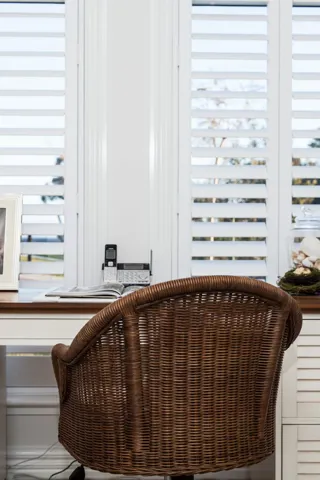 Office room with a brown wicker chair and cream and brown desk complimented by open white shutters