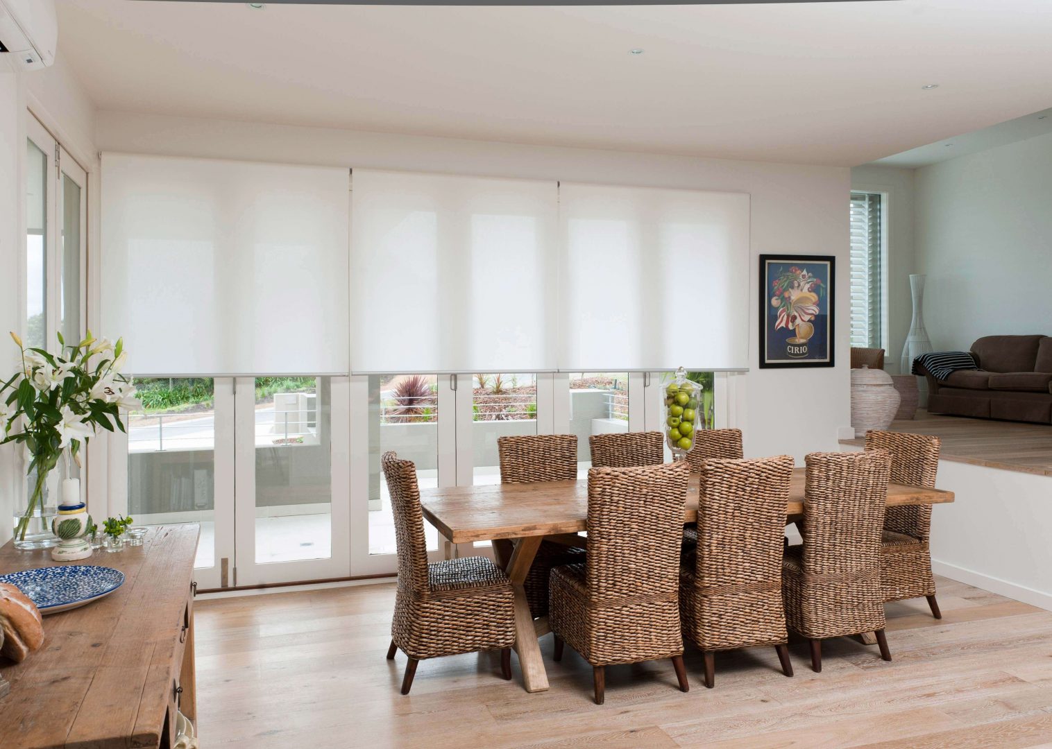 Dining room with brown wicker dining chairs and timber table complimented by a white sheer roller blind