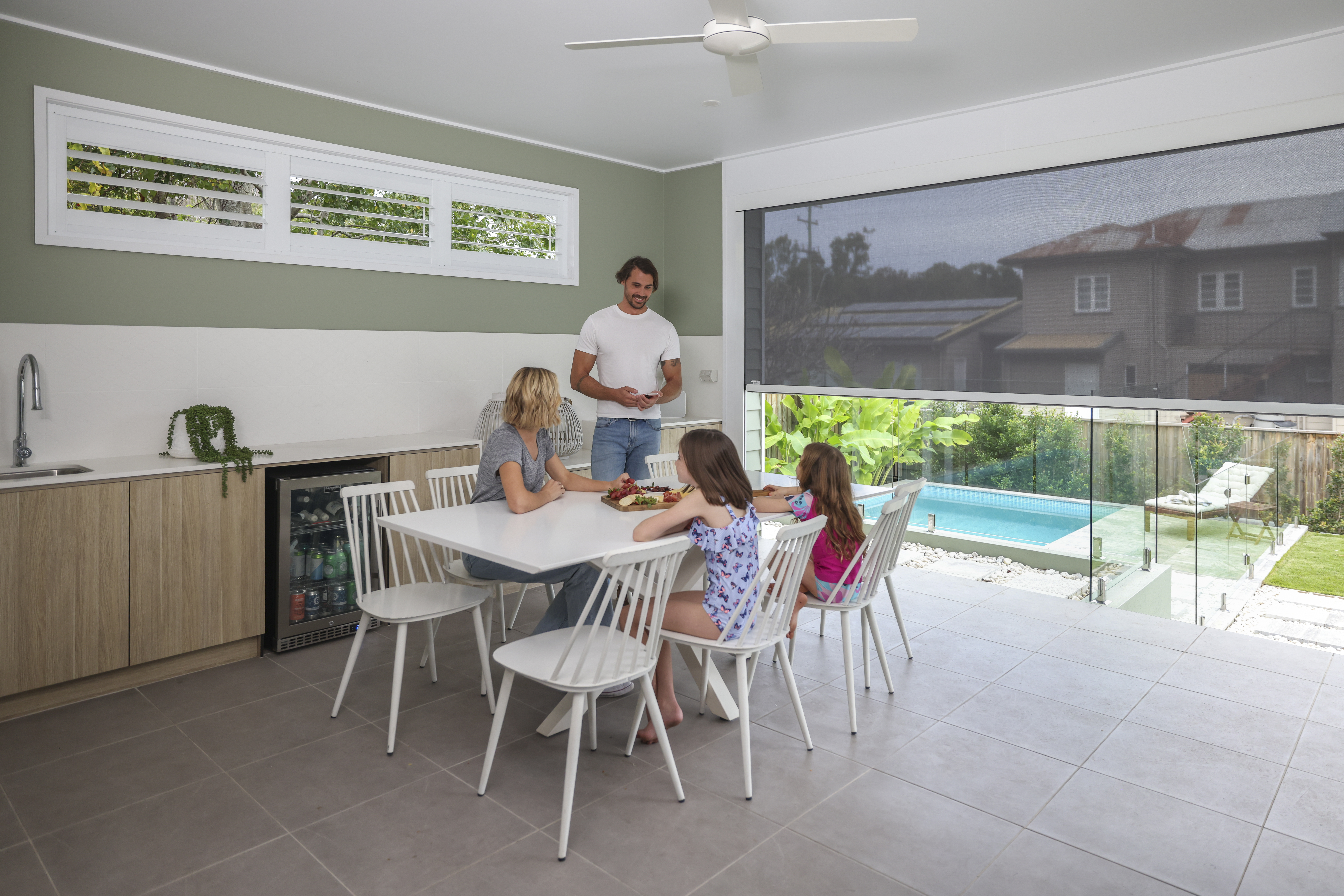 Dark grey awning half open to a family dining in an entertaining space with a sage green wall and white and timber furniture