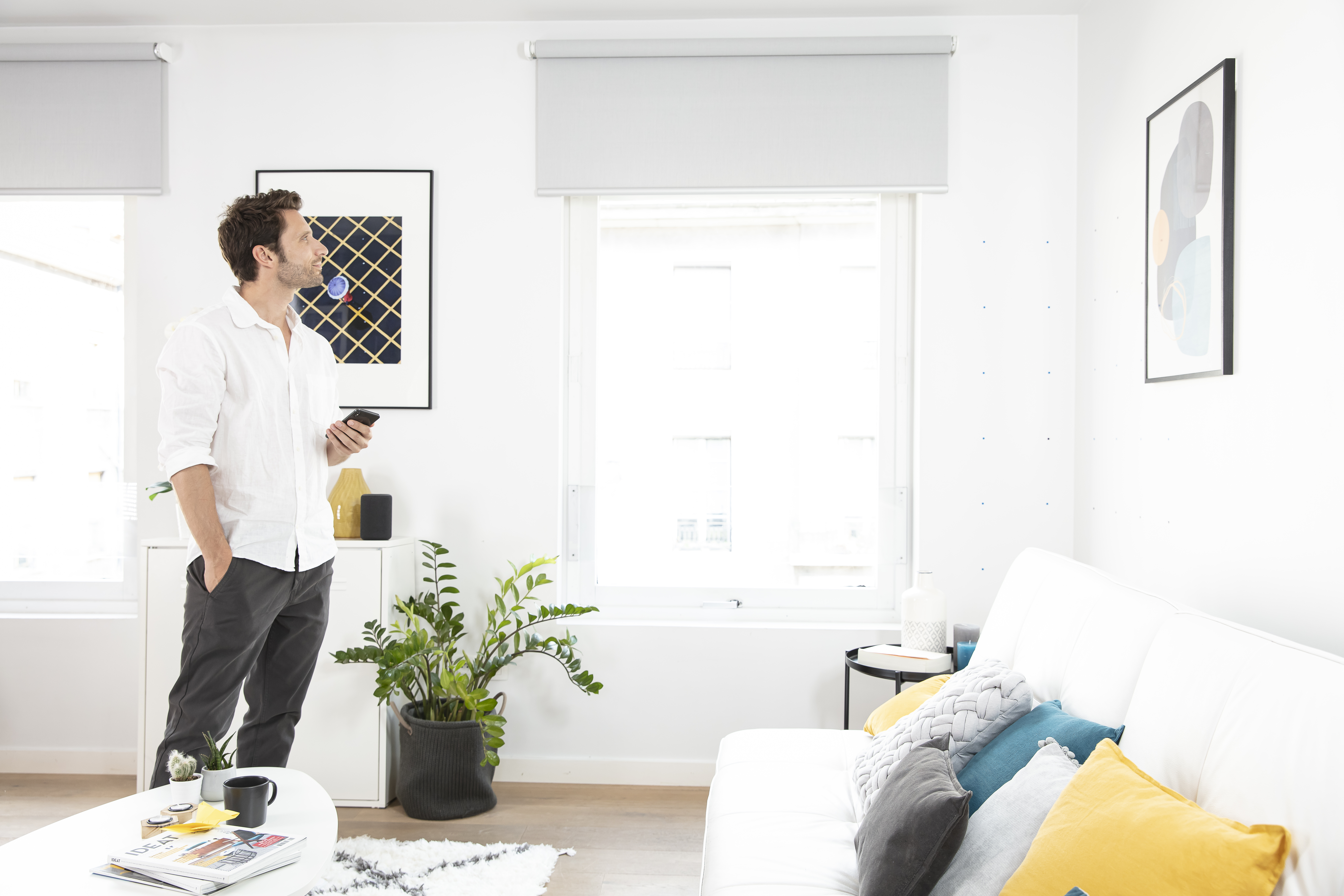 A man standing with a remote in hand looking at white dual roller blinds in a white living room