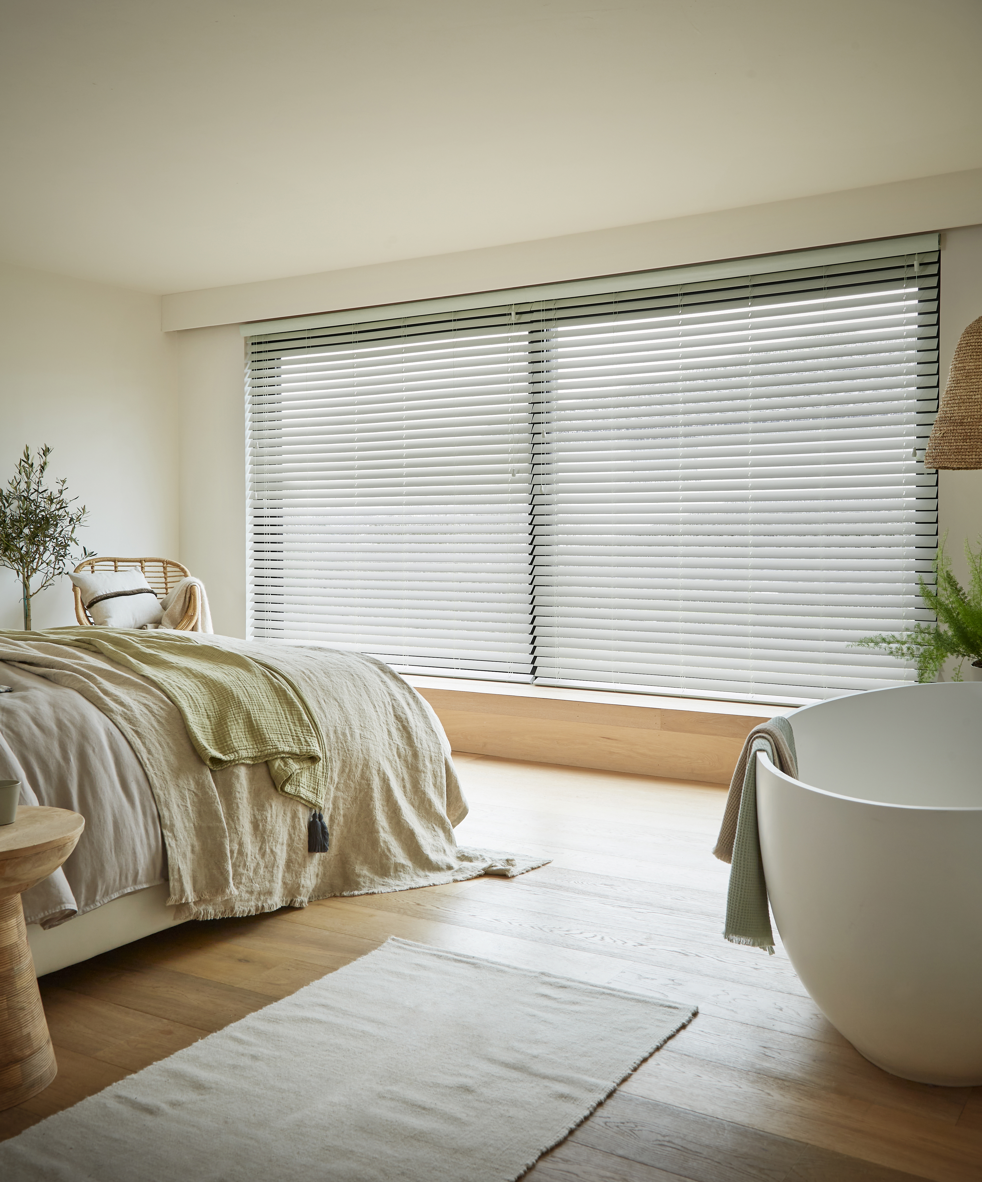 Calming bedroom with large windows covered by light grey Venetian blinds, a cosy bed with soft linens, a freestanding bathtub, and natural wooden accents for a serene atmosphere