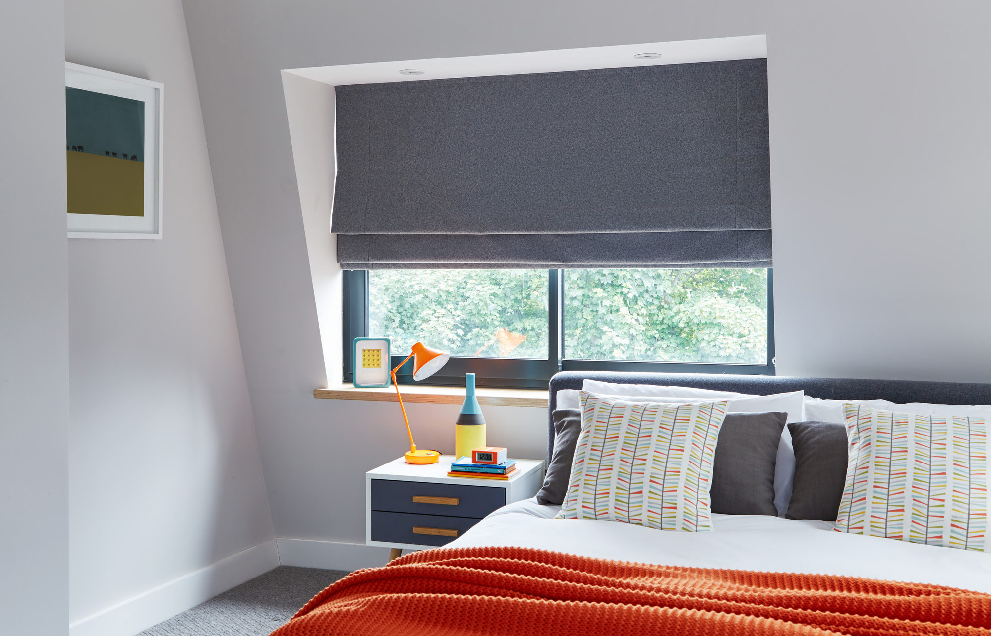 Grey and white toned bedroom with bright orange throw blanket and a dark grey roman blind