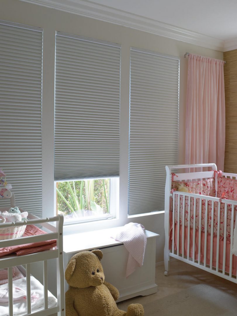 Children's bedroom featuring white cellular blinds, pink curtains, two white cribs, and a large teddy bear on the floor
