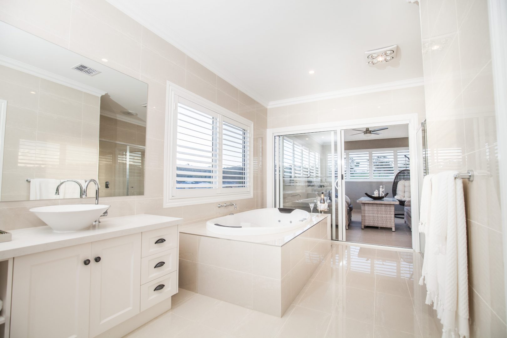 White PVC shutters open in cream tiled bathroom with white sink and bathtub