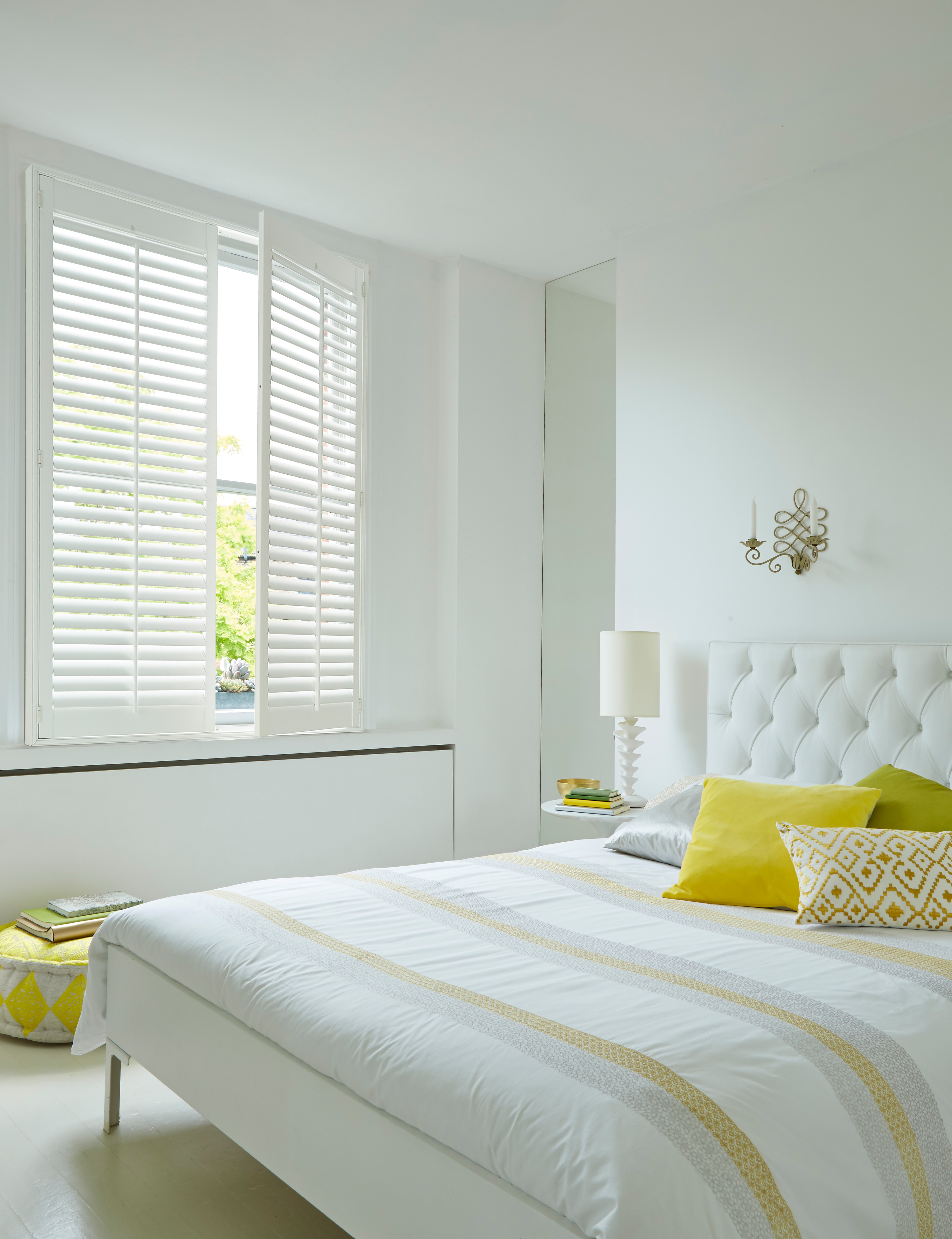 Bright bedroom featuring white plantation shutters, a tufted white bed with yellow accent pillows, and minimalist decor for a clean, modern aesthetic