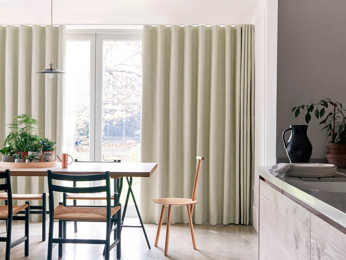 Bright dining area with floor to ceiling beige curtains, a wooden dining table surrounded by black chairs, and various potted plants