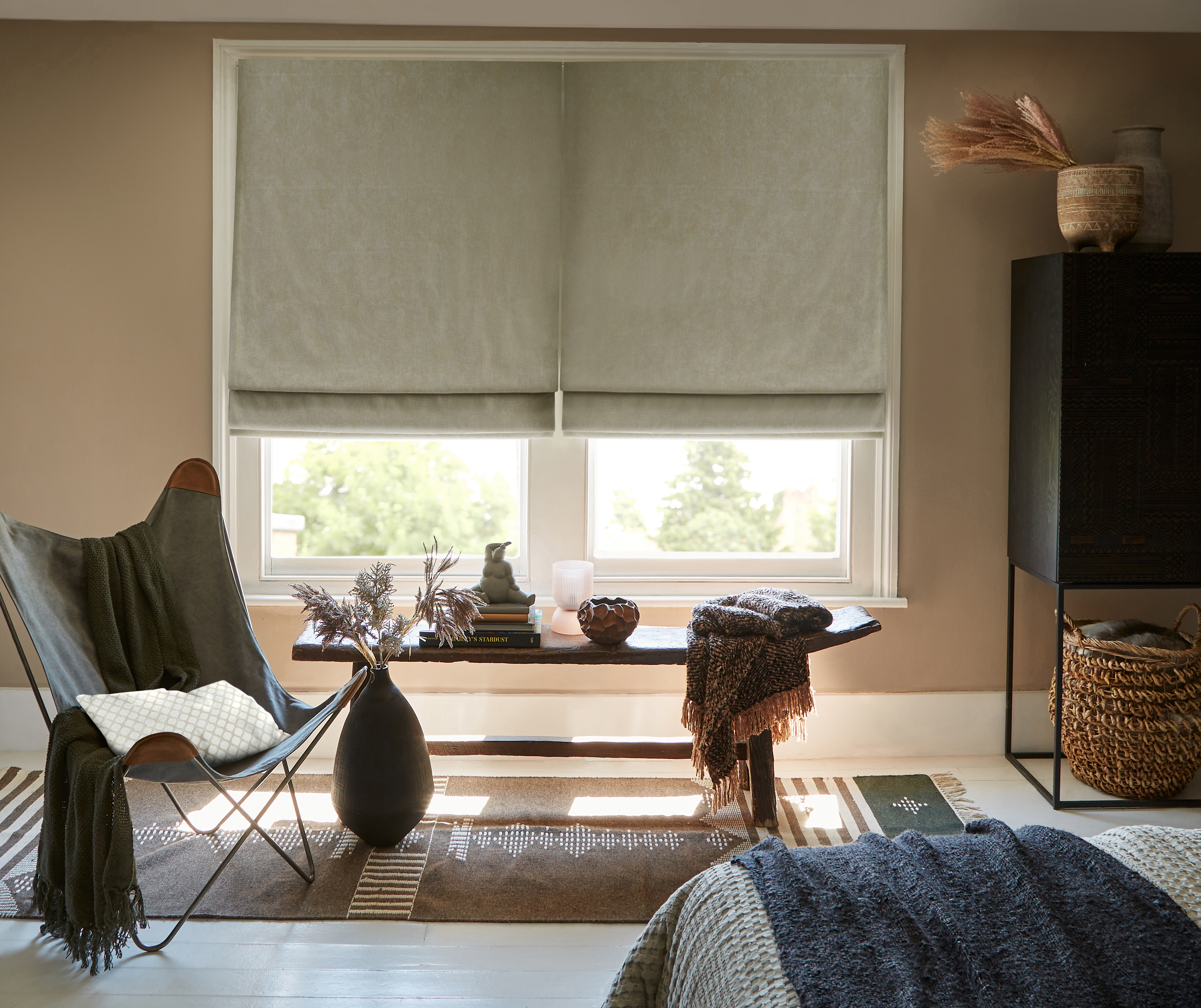 Cosy bedroom with soft grey Roman blinds, a rustic wooden bench, a leather butterfly chair, and woven accents, creating a warm and inviting space with natural decor elements