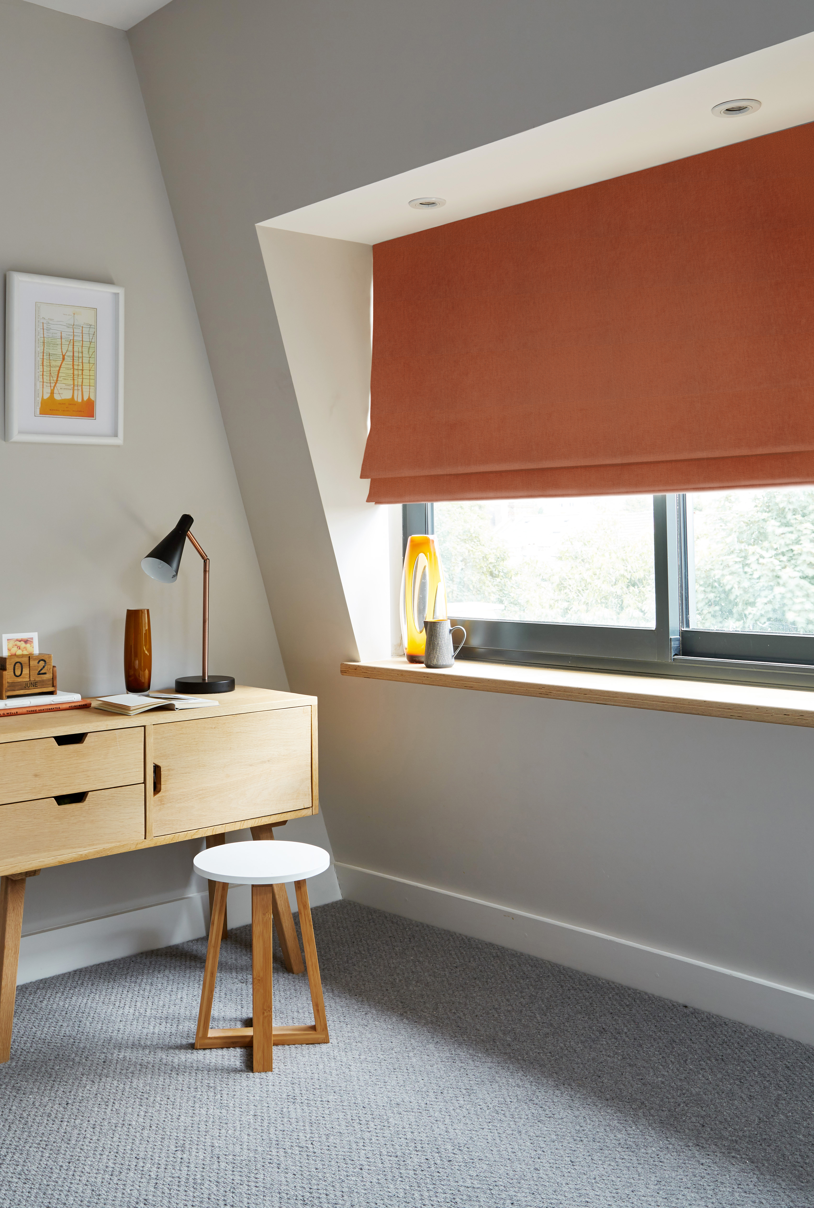 Bright workspace with a burnt orange Roman blind, a wooden desk with a modern lamp, and a small stool, creating a warm and inviting home office