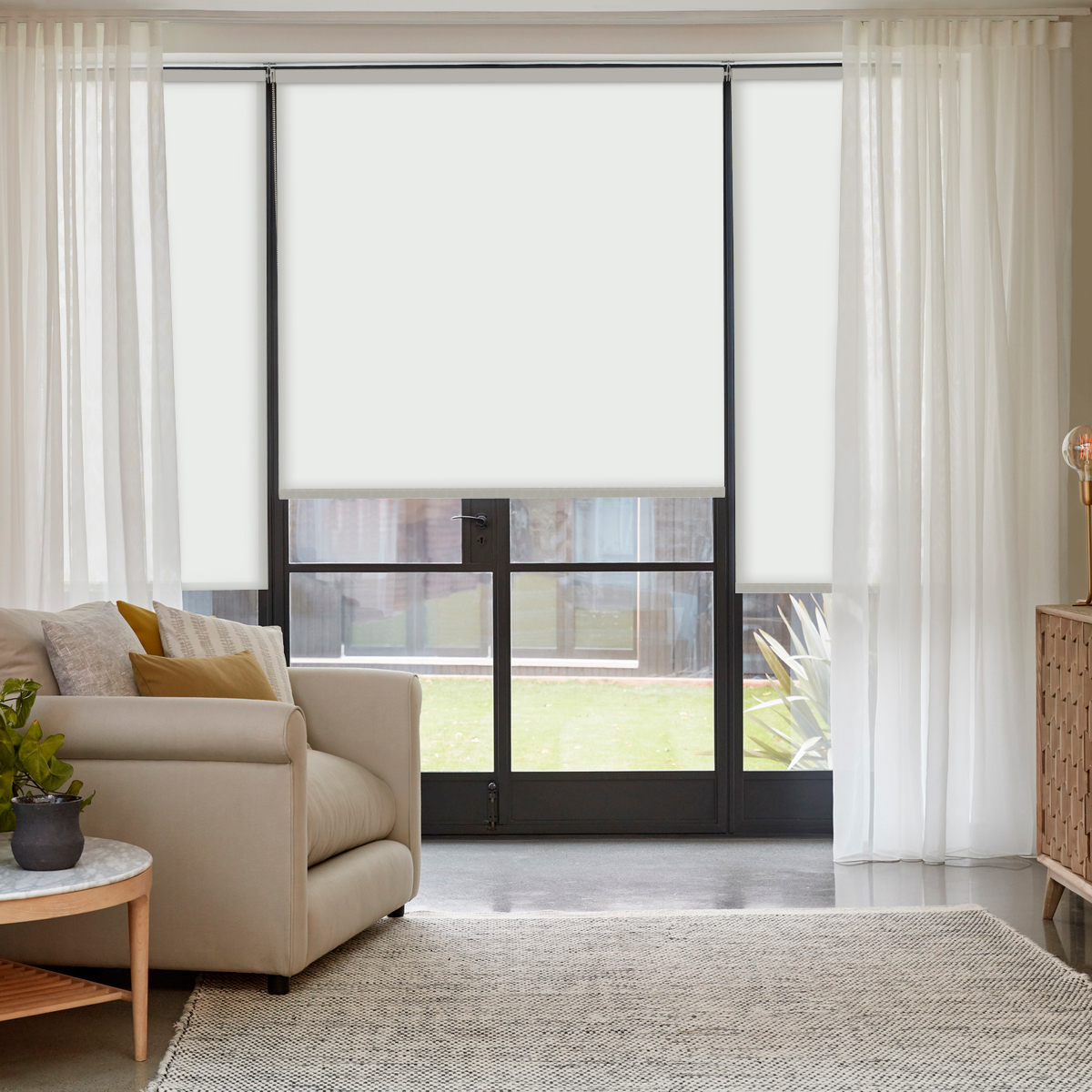 Bright living room featuring sheer voile curtains layered with roller blinds, a beige sofa with accent pillows, a wooden side table, and a neutral-toned rug