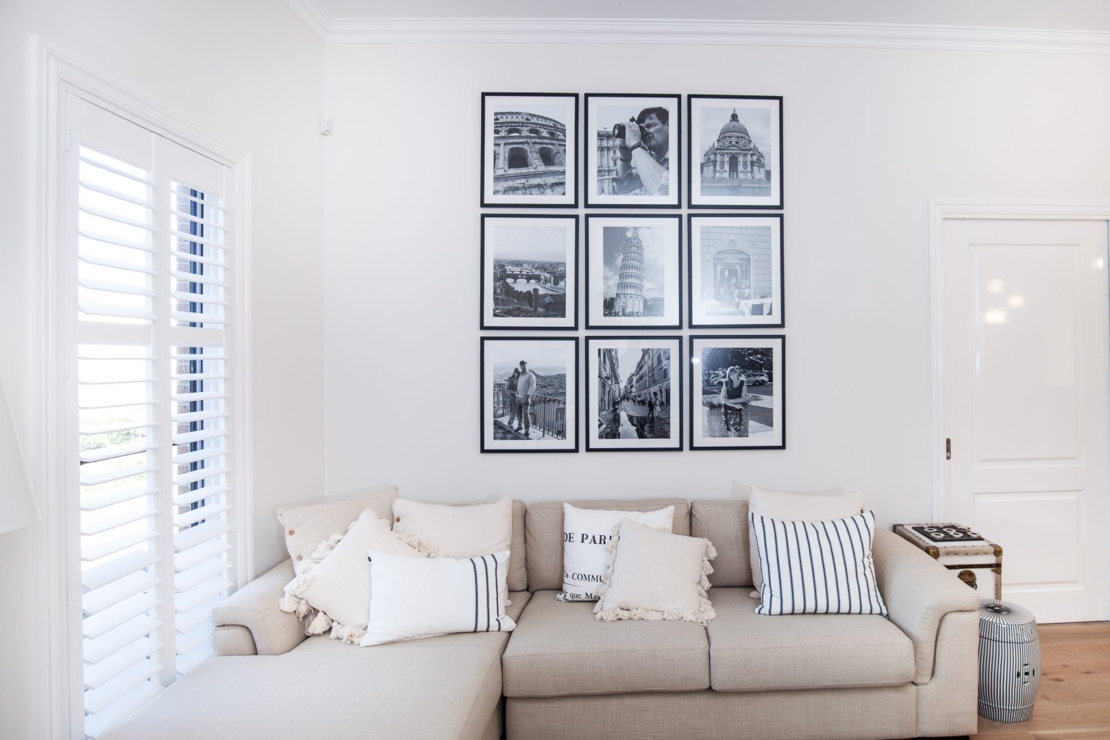 White PVC shutters semi open in white walled living room with black and white pictures and cream couch