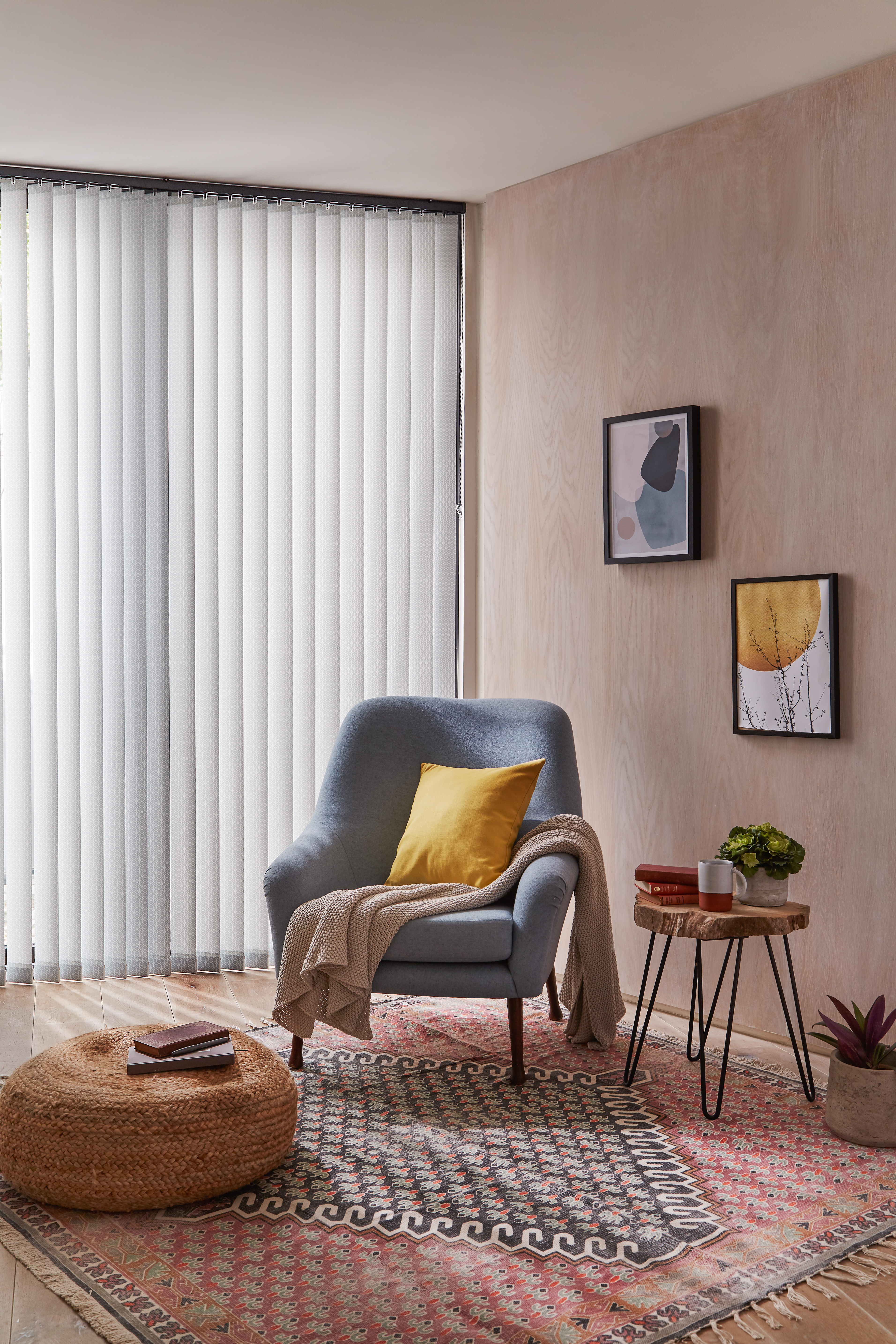 Cosy reading nook with light grey vertical blinds, a blue armchair accented by a yellow cushion, a woven pouf, and a patterned rug, creating a warm and inviting space