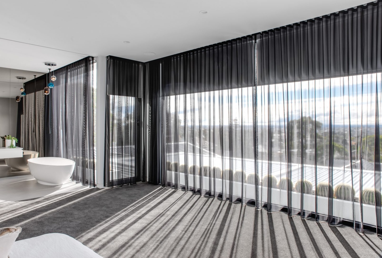 A large open space with grey carpet, a white bathtub and closed black sheer curtains with a dark grey roller blind