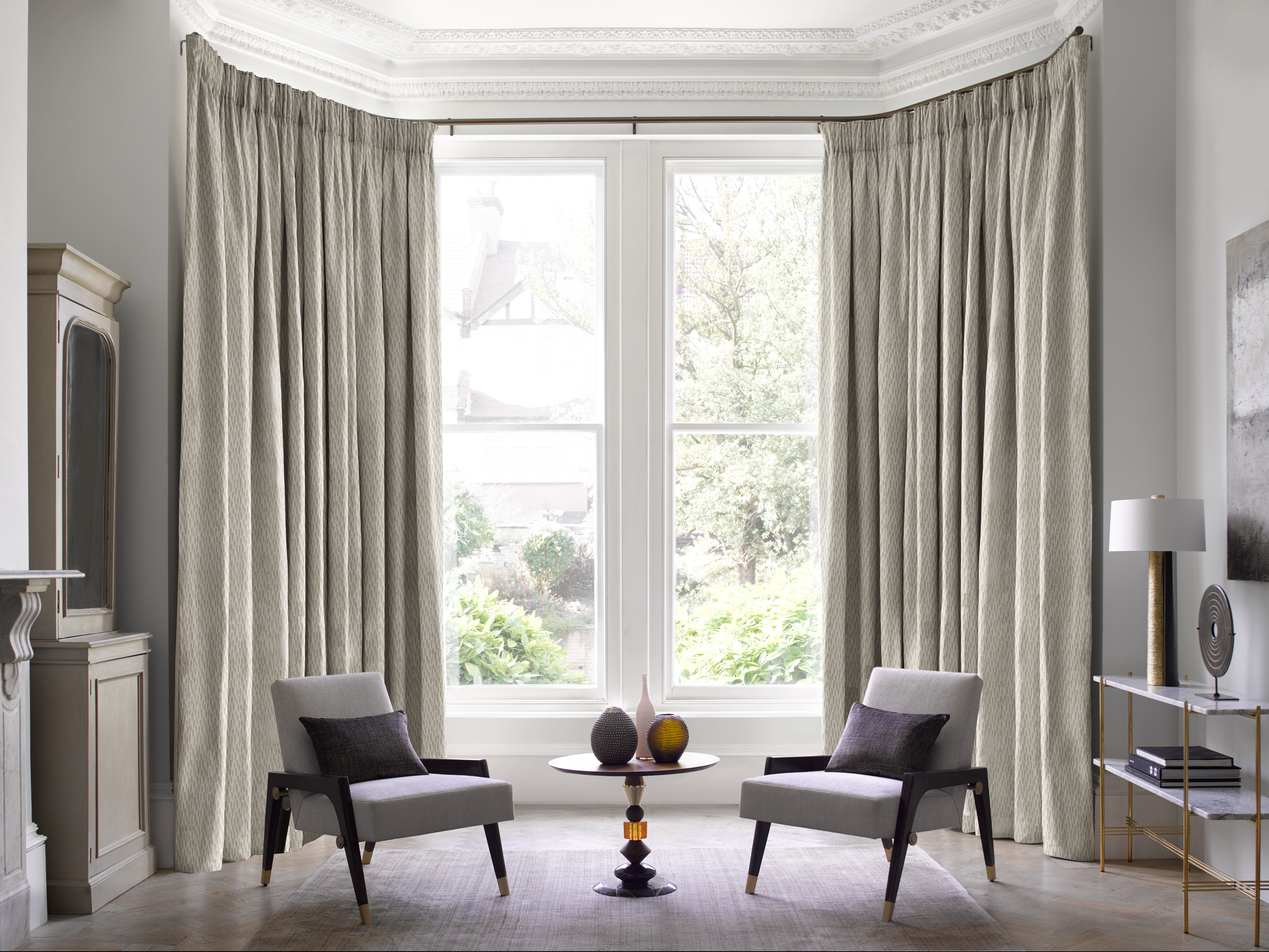 Elegant living room featuring tall windows framed by floor-to-ceiling light grey pencil pleat curtains, two modern armchairs, and a small round table